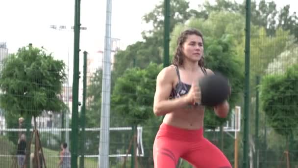 sport and healthy lifestyle. Young caucasian woman curly hair with tattoo. Athlete doing exercise training leg muscles and buttocks. Girl squats and jumps with ball in hands at city workout stadium - Metraje, vídeo