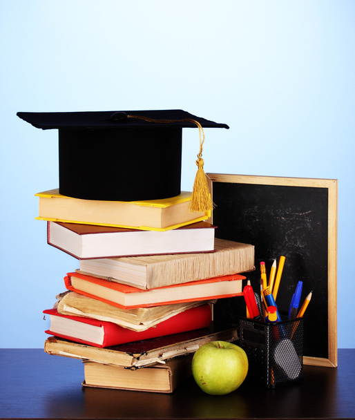 Books and magister cap against school board on wooden table on blue background - Foto, Bild