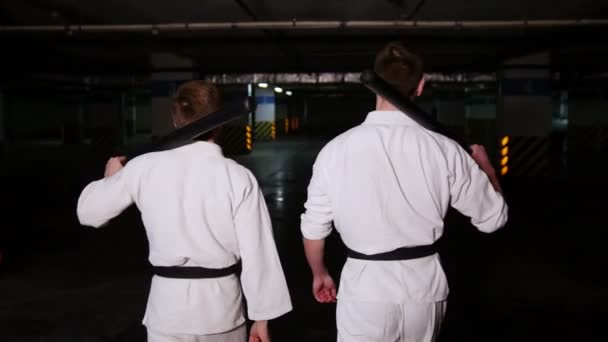 Dois homens em quimono andando em um estacionamento segurando uma espada de treinamento em seus ombros
 - Filmagem, Vídeo