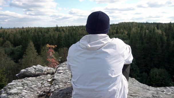 Sám muž sedí na vrcholu vysoké hory při pohledu na zatažené oblohy a stromy vrcholy. Pohled zezadu na muže v klobouku a bílá bunda sedí na skále, při pohledu na krásný les. - Fotografie, Obrázek