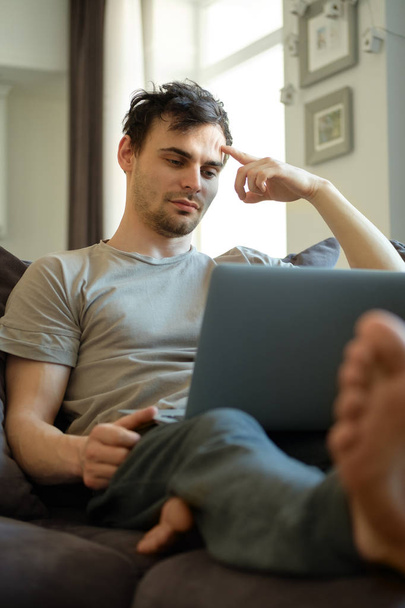 Millennial man lying on sofa with laptop computer spending time for messaging with followers from social networks, young male spending his leisure time in modern home interior with stylish furniture - Foto, Imagem