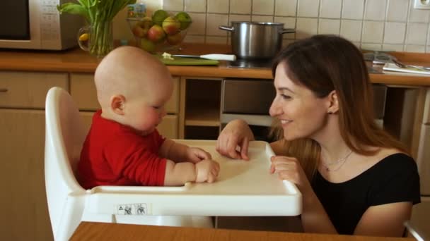 Mother feeding hungry baby in highchair in kitchen. The woman gently smiles at the baby. Mothers Day - Video