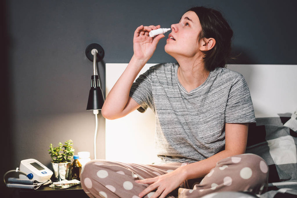 Тема сезонная холодная, насморк инфекции вируса гриппа носа. Молодая кавказская женщина на кровати в своей спальне использует капли лекарств в носу для соплей. Аллергический ренит и синусит
 - Фото, изображение