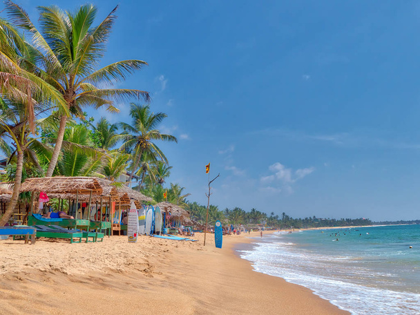 pláži scenérie v Hikkaduwa Beach se spoustou kokosové palmy, pláže s jemným pískem a Palmový list chaty na fascinující tropické ostrov Srí Lanka v Indickém oceánu - Fotografie, Obrázek