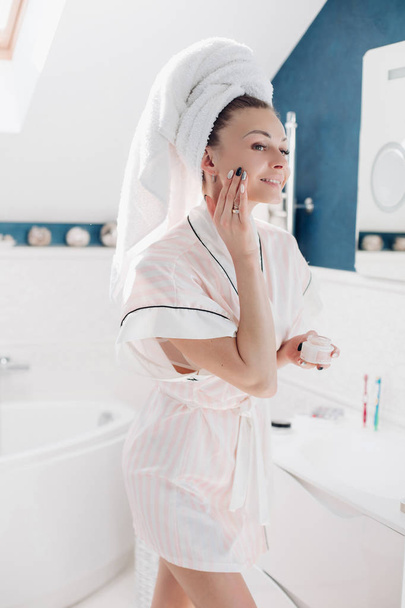 Beautiful young woman applying cream on her face in bathroom. - Fotoğraf, Görsel