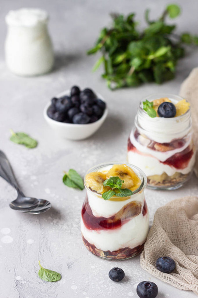 Granola, blueberry and banana greek yogurt parfaits in mason jars. Healthy breakfast. A light grey concrete background. Copy space. - Фото, изображение