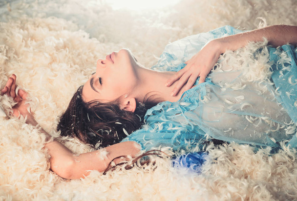 Woman with long hair in tender pajama relaxing. Sweet dreams concept. Lady in transparent blue nightie lay on bed and sleep. Girl on calm face lay on bed covered with feathers and fluff. - Photo, Image
