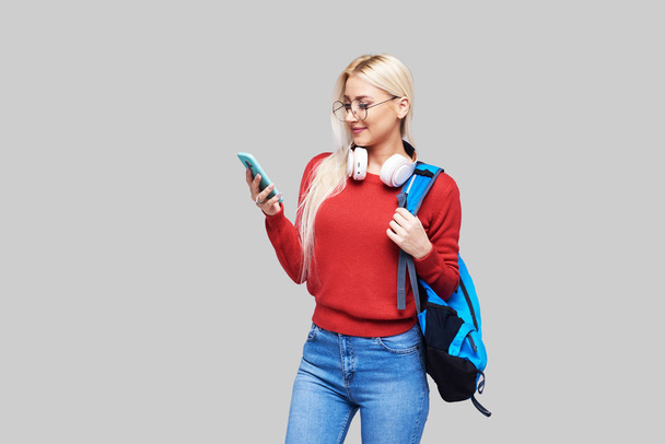 Portrait of a cheerful happy blond female student with blue backpack listening online lecture on phone with headphones isolated over grey background. Copy space for text on the left side - Photo, image