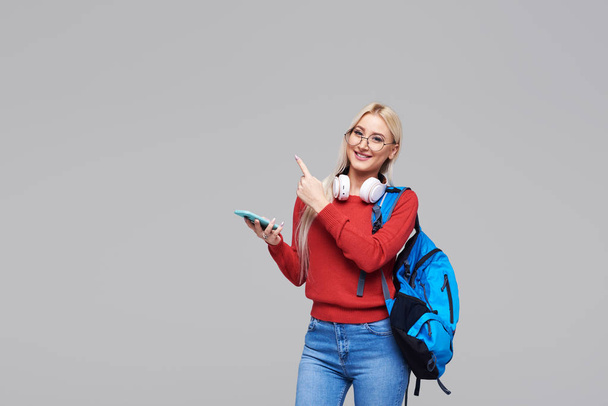 Portrait of a cheerful happy blond female student with blue backpack listening online lecture on phone with headphones isolated over grey background. Copy space for text on the left side - Фото, изображение