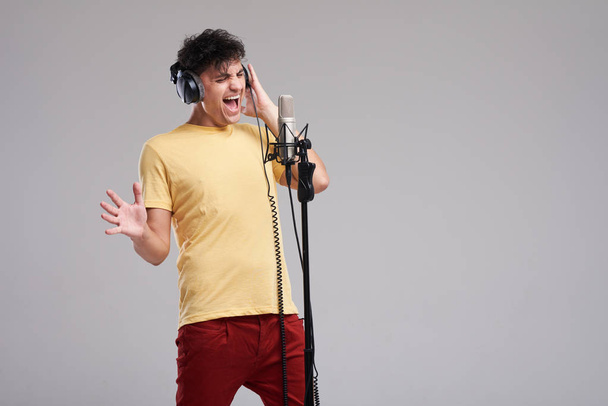 Portret mężczyzny piosenkarz rockowy w profesjonalne słuchawki studyjne, utrzymywanie statycznej mic, głośno śpiewa piosenkę na szarym tle. Pojęcie muzyki rockowej i rave - Zdjęcie, obraz