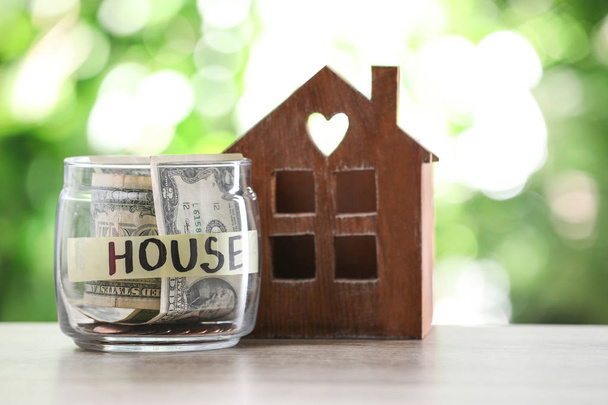 Glass jar with money for mortgage and house model on table against blurred background - Foto, afbeelding