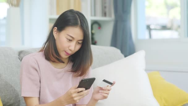 Дівчина, яка посміхається азіатській жінці, користується смартфоном і купує онлайн за кредитною карткою, лежачи на дивані, коли відпочиває вдома. Стиль життя латин і гізпанічна етнічна приналежність Жінки вдома концепція. - Кадри, відео