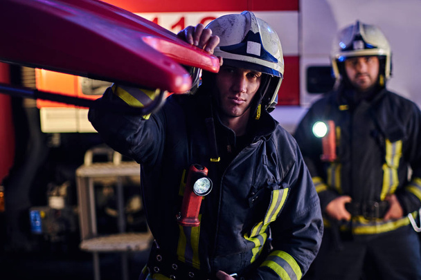 Двое храбрых пожарных в защитной форме стоят рядом с пожарной машиной. Прибытие по вызову в ночное время
 - Фото, изображение