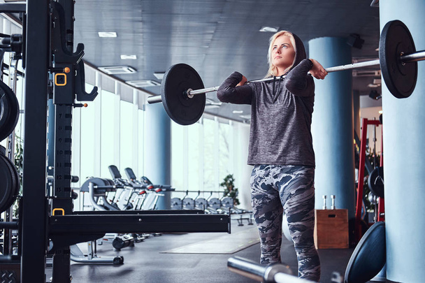 Beautiful sportive girl wearing hoodie doing squats with barbell in the modern gym - Photo, Image
