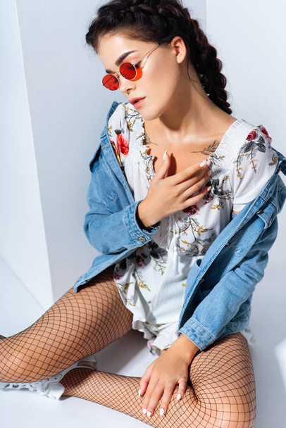 stylish woman with braid in sunglasses and jacket with white ethnic embroidery sitting on white floor  - Fotografie, Obrázek