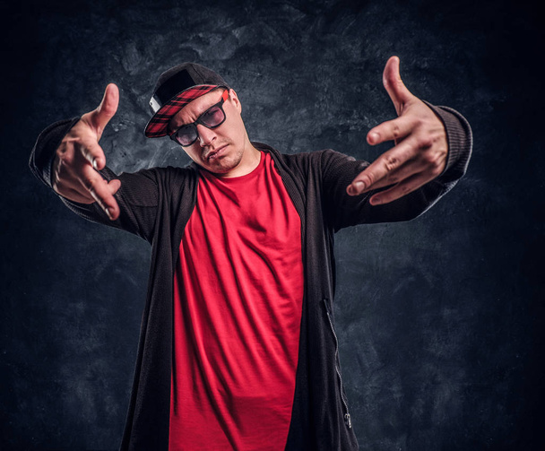 Portrait of a young rapper dressed in a hip-hop style, posing for a camera. Studio photo against a dark wall - Photo, Image