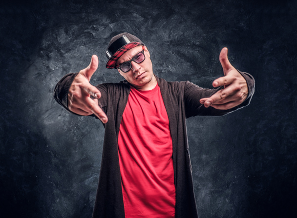 Πορτρέτο του ένας νεαρός ράπερ ντυμένη σε στυλ hip-hop, ποζάρει για μια φωτογραφική μηχανή. Στούντιο φωτογραφία ενάντια σε έναν τοίχο σκούρο - Φωτογραφία, εικόνα
