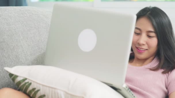 Молодая деловая внештатная азиатка, работающая на ноутбуке, проверяя социальные сети, лежа на диване, когда отдыхает в гостиной дома. Стиль жизни латинские и латиноамериканские этнические женщины в доме концепции. - Кадры, видео