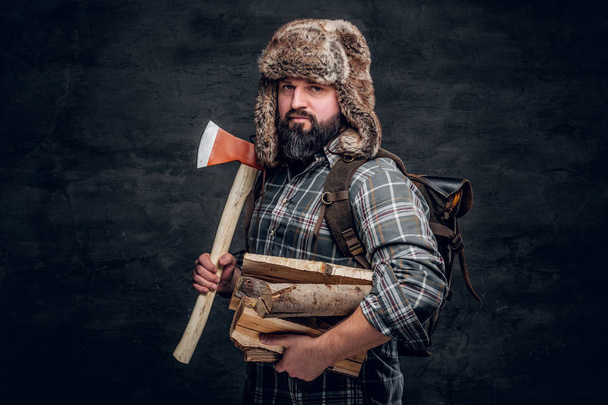 Portrait d'un bûcheron barbu avec un sac à dos habillé d'une chemise à carreaux et d'un chapeau de trappeur tenant du bois de chauffage et de la hache
. - Photo, image