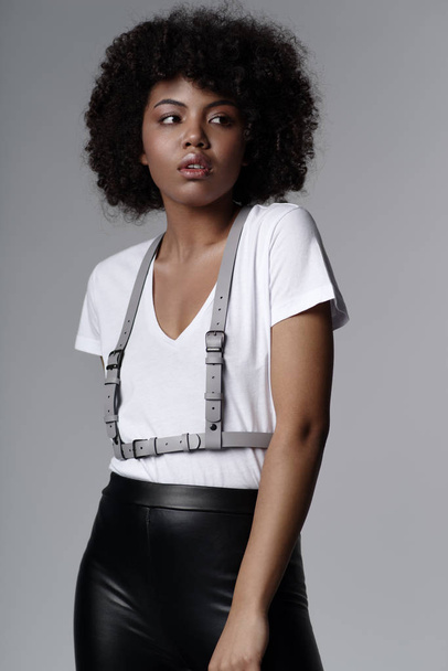 beautiful african american woman with curly hairstyle wearing stylish clothes and leather female sword belt and posing on light grey background  - Fotó, kép