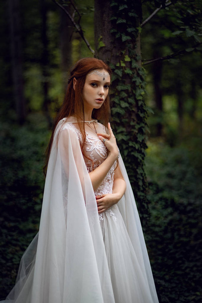 attractive ginger elf woman in white dress and cape posing in green garden  - Fotó, kép