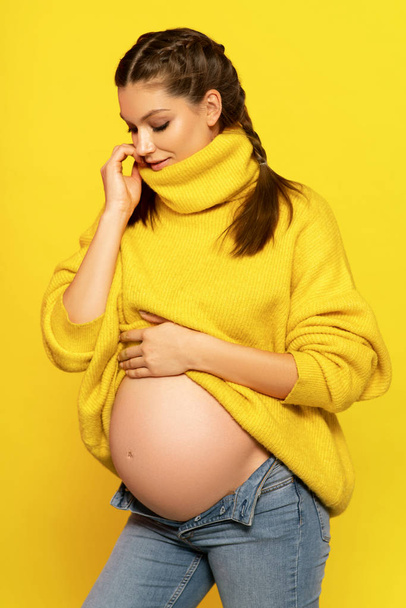 attractive pregnant woman with braids posing in warm sweater on bright yellow background - Fotó, kép