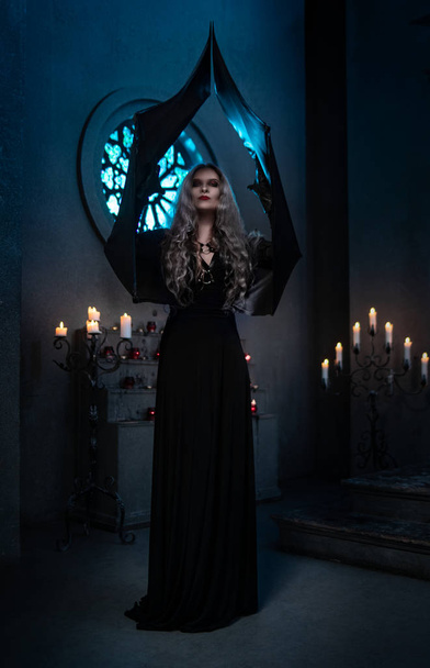 spooky woman in witch costume posing with raised arms in dark gothic building with candles on background - Fotoğraf, Görsel