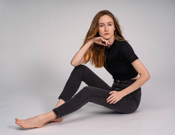 beautiful ginger woman in dark clothes posing while sitting on grey studio background - Fotó, kép