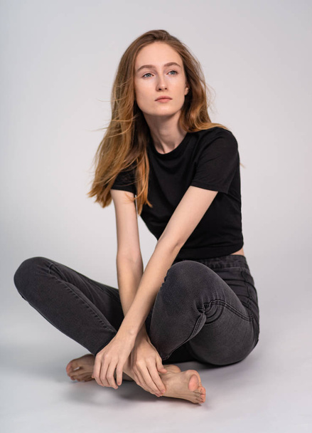 hermosa mujer de jengibre en ropa oscura posando mientras está sentado con las piernas cruzadas sobre fondo gris estudio
 - Foto, imagen