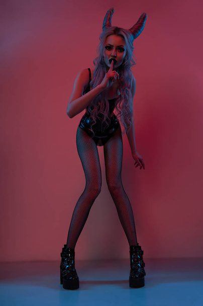 attractive blonde woman with devil horns wearing black latex body posing on dark pink background - Foto, afbeelding