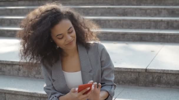 Beautiful young girl with dark curly hair using her cell phone, outdoor. - Felvétel, videó
