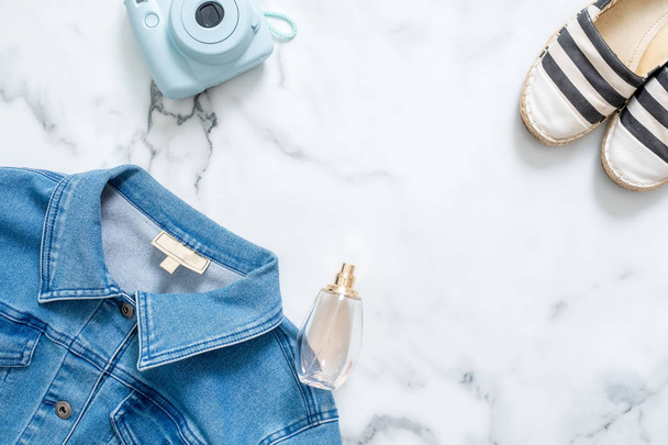 Beauty blogger desk with feminine jeans jacket, hipster instant photo camera,  fragrant bottle of perfume, fashionable striped sandals. Casual women's clothing on marble background on flat lay style - Foto, imagen