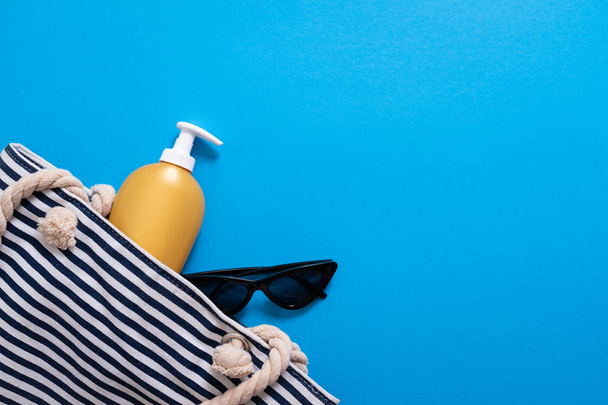 Summer composition. Stripped beach bag with suntan cream and sunglasses. Summer holiday concept, sunscreen cosmetic and accessories on blue sea background. - Photo, image