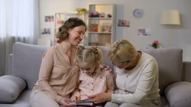 Granny and mother showing photo album pictures to small girl, family history - Materiał filmowy, wideo