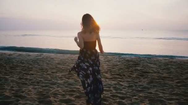 Camera volgt gelukkig opgewonden vrouwelijke reiziger in mooie jurk loopt naar de komende golf van de Oceaan om te kijken naar de zonsondergang op het strand. - Video