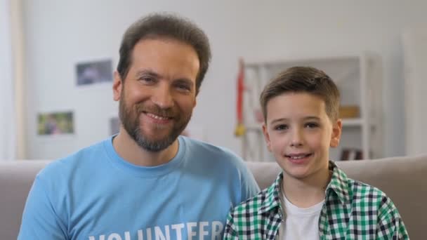 Cheerful little boy and adult man volunteer showing thumbs-up into camera - Materiał filmowy, wideo