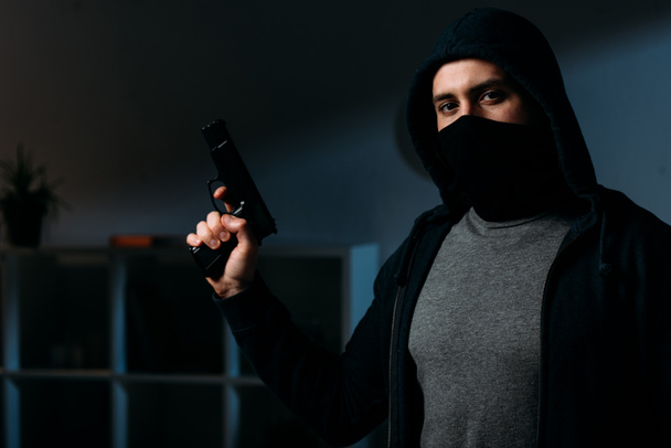 Robber in mask holding gun and looking at camera in dark room - Zdjęcie, obraz