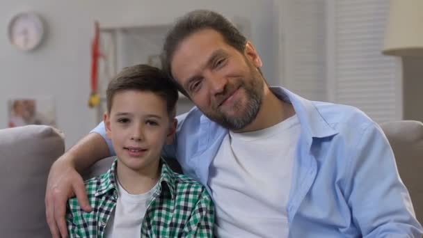 Father hugging son, sitting on sofa at home and looking at camera, happy family - Materiał filmowy, wideo