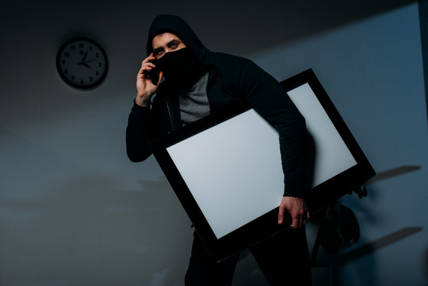 Thief in mask talking on smartphone while stealing flat-screen tv with blank screen - Photo, Image