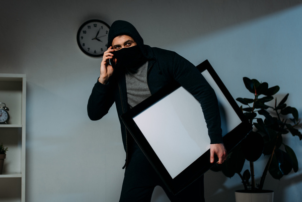 Ladrão em máscara falando no smartphone enquanto rouba tv de tela plana com tela em branco
 - Foto, Imagem