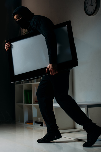 Cautious thief in mask stealing flat-screen tv with blank screen - Fotoğraf, Görsel