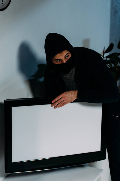 Sospechoso ladrón en máscara robando televisión de pantalla plana con pantalla en blanco
 - Foto, imagen