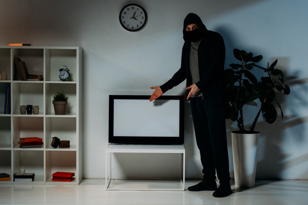 Ladrão de máscara e capuz apontando com as mãos para a televisão de tela plana com tela em branco
 - Foto, Imagem