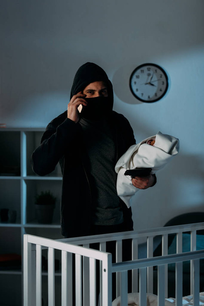 Kidnapper in mask holding infant child and talking on smartphone - Photo, Image
