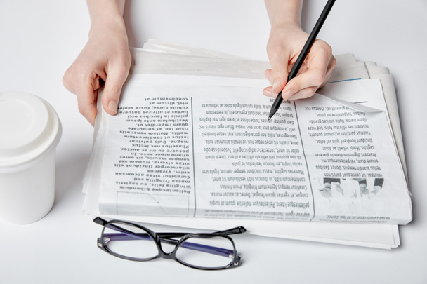 cropped view of woman holding pencil near newspaper, glasses and paper cup with drink on white  - Фото, изображение
