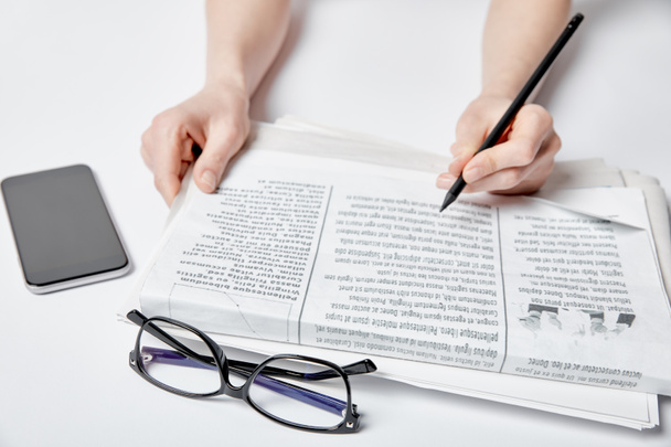 cropped view of woman holding pencil near newspaper, glasses and smartphone with blank screen on white  - Fotó, kép