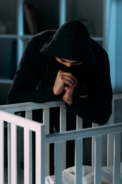 Kidnapper in black hoodie looking in crib and showing please gesture - Foto, immagini