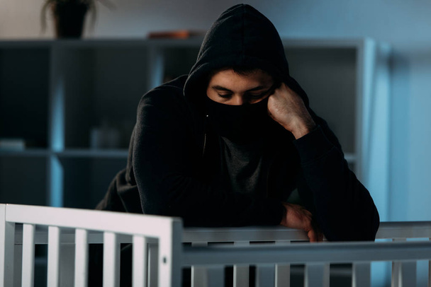 Pensive kidnapper in black mask and hoodie looking in crib - Фото, изображение