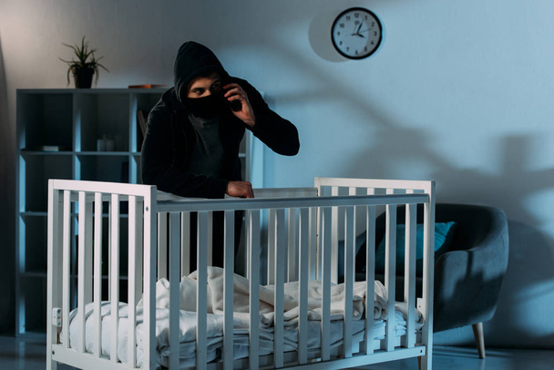 Kidnapper in black mask standing near crib and talking on smartphone - Photo, Image