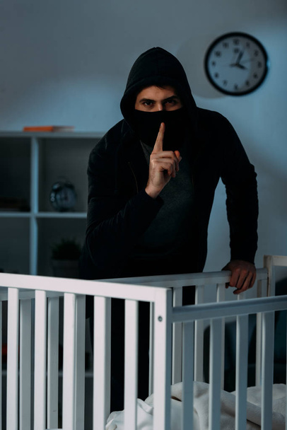 Kidnapper in mask standing near crib and showing hush sign - Valokuva, kuva